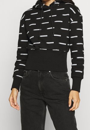 Hoodie nero corto con motivi logo bianchi, cappuccio con cordino e polsini e orlo a coste. Abbinato a jeans scuri, che mostrano uno stile casual.