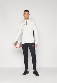 Vit långärmad topp med svarta och röda detaljer, svarta träningbyxor och svarta fotbollsskor. Håller i en mönstrad fotboll.