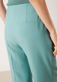 Ljus turkosa skräddarsydda byxor gjorda av ett slätt tyg, med en bakficka och en strömlinjeformad siluett. Närbild av sidan och baksidan.
