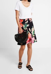 T-shirt blanc à col en V, jupe noire à fleurs avec motifs roses et verts, sac rond noir, et sandales noires à enfiler. Tenue décontractée.