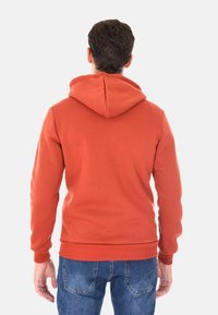 Felpa rossa con orlo e polsini a coste, dotata di un grande cappuccio. Il tessuto sembra morbido e con una texture, indossata con jeans blu.