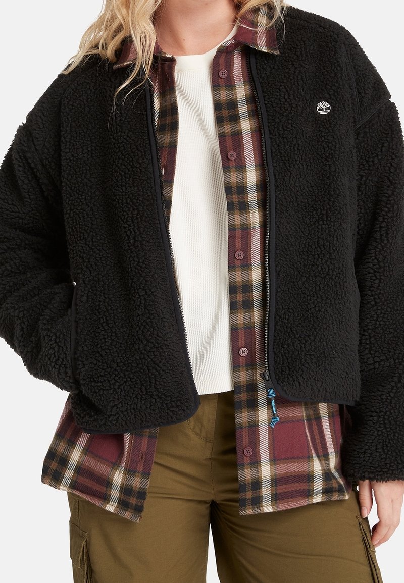 Czarna kurtka polarowa o fakturowanym wykończeniu, z zamkiem błyskawicznym i bocznymi kieszeniami, nałożona na koszulę flanelową w kratę w kolorze bordowym i beżowym.