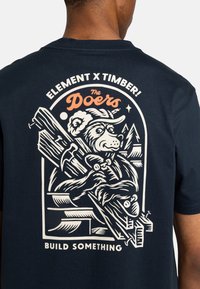 Navyblaues T-Shirt mit einem Grafikdesign eines Bären in einem Hut, der Werkzeuge hält, mit weißem und orangefarbenem Text "ELEMENT X TIMBER!" und "BAUET ETWAS."