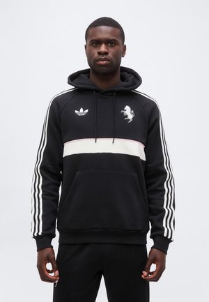 Junger Mann trägt einen schwarzen Hoodie mit weißem horizontalem Streifen, drei weißen Streifen auf den Ärmeln und zwei Logos auf der Brust, steht vor grauem Hintergrund.