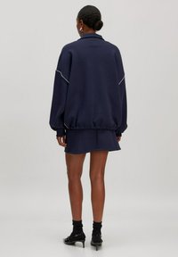 Navyblauwe oversized pullover met witte piping details, gecombineerd met een bijpassende minirok, gemaakt van gladde stof, weergegeven vanuit de achterkant.