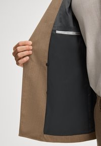 Brauner Blazer mit einem strukturierten Außenmaterial. Das Innenfutter verfügt über ein glattes schwarzes Futter und ein gestreiftes Taschendetail. Zwei sichtbare Knöpfe.
