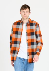 Rutet skjorte i orange, grå og sort; knaplukning, lange ærmer og et struktureret stof. Båret over en hvid T-shirt med blå jeans.