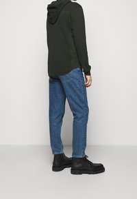 Sudadera con capucha verde oscuro de tejido texturizado, combinada con jeans de mezclilla azul y botas de tobillo negras con cordones. Ajuste ligeramente relajado.