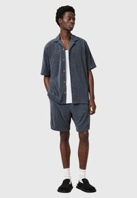 Mannelijk two-piece outfit in donkergrijze terry stof, bestaande uit een shirt met korte mouwen en knopen en bijpassende shorts, gecombineerd met zwarte instappers.