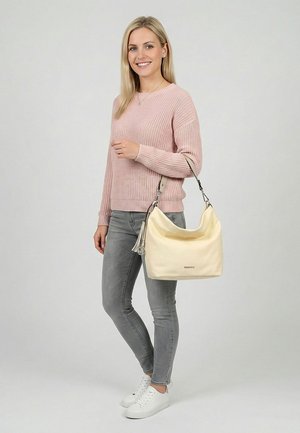 Joven mujer con suéter rosa y vaqueros grises sosteniendo un bolso beige con borla, de pie frente a un fondo liso gris claro, sonriendo.