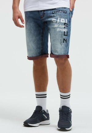 Mann trägt hochgekrempelte blaue Jeansshorts mit aufgedrucktem Text, weiße Socken mit schwarzen Streifen und dunkelblaue Turnschuhe.