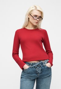 Νεαρή γυναίκα με μακριά ξανθά μαλλιά, που φοράει μαύρα γυαλιά, κόκκινο μακρυμάνικο τοπ και μπλε τζιν με κορδόνια, με τα χέρια στις τσέπες.