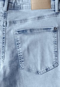 Un tissu en denim bleu clair présente une poche arrière avec un rabat pointu, un léger dégradé et des coutures visibles. Une étiquette en cuir tawny est attachée.