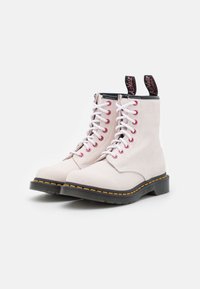 Light pink suede combat boots with white laces, red eyelets, black accents, and distinctive yellow stitching on the black sole.