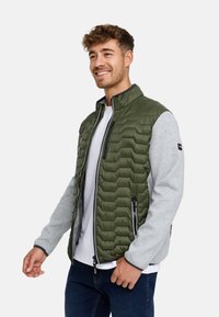 Ung mand med kort krøllet hår, smilende, iført grøn quiltet vest med grå ærmer, hvid skjorte og blå jeans, stående mod en hvid baggrund.