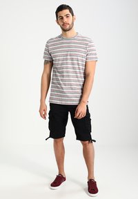 T-shirt grigio a righe con linee rosse e blu, abbinato a pantaloni cargo neri con tasche laterali. Sneakers rosse completano l'outfit.