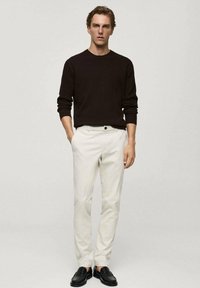 Ciemnobrązowy sweter w prążki, białe spodnie i czarne buty. Model stoi prosto, prezentując czyste linie i neutralną paletę kolorów.
