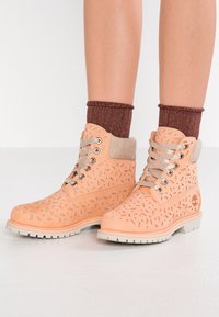 Stivaletti alla caviglia in pelle pesca con design perforato, dettagli argento e lacci grigi. Suola bianca in gomma e calze lavorate marroni visibili.
