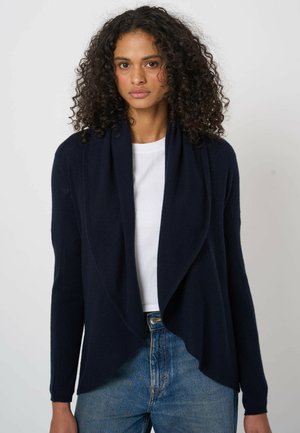 Kobieta o kręconych włosach, ubrana w ciemnogranatowy otwarty kardigan na białej koszuli i niebieskich dżinsach, stojąca na jednolitym tle.