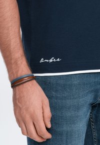 Námořnické polo tričko s bílými detaily, na lemu je vyšívané logo "Ombre". Na ruce ozdobené koženými a kovovými náramky. Viditelné džíny.