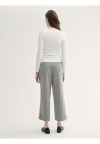 Witte lange mouw fitted top met ribbelstructuur, gecombineerd met grijze broek met brede pijpen in herringbone dessin en zwarte platte schoenen.