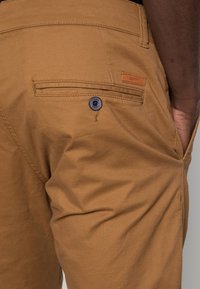 Bermudas de algodón marrón con cierre de botón, bolsillo trasero y etiqueta de marca cosida. Textura suave con un corte clásico.