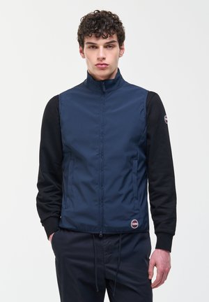 Jeune homme portant un gilet zippé bleu marine par-dessus un t-shirt à manches longues noir, debout devant un fond uni clair.