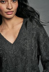 Grå strikket sweater med V-hals, der har et mønstret design og små, skinnende pynt langs siderne. Blød og hyggelig udseende.