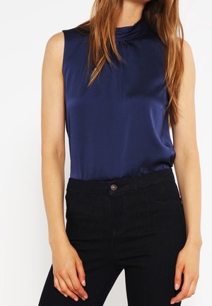 Blusa sin mangas de satén azul marino combinada con pantalones negros de cintura alta en una persona de pie con cabello largo y castaño.