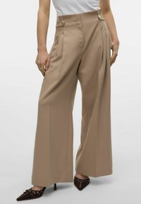 Beige, høj talje bukser med plisseringer, to sidelukninger og vidde i benene. Stoffet har en glat tekstur. Bærer med mørke spidse hæle.