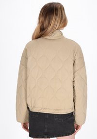 Beige gesteppte Jacke mit hohem Kragen, versehen mit einem gewellten gestickten Muster und elastischem Saum. Das Modell trägt einen schwarzen Jeansrock.