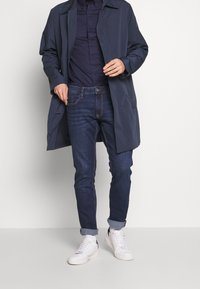 Uomo che indossa un cappotto blu scuro, camicia blu navy, jeans scuri arrotolati e sneakers bianche, in piedi contro uno sfondo chiaro uniforme.