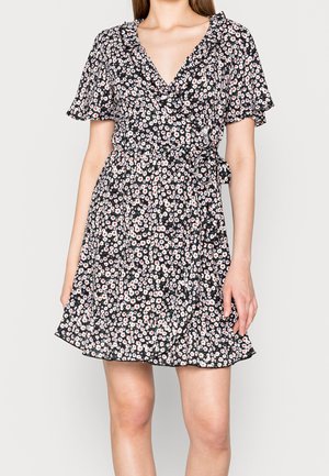 Mujer que lleva un vestido cruzado negro con estampado floral, mangas cortas con volantes y cinturón atado a la cintura.