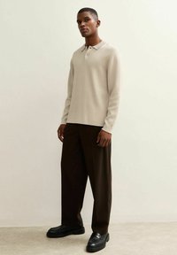 Beige poloshirt met lange mouwen en een kraag, gecombineerd met bruin wijde pantalon en zwarte schoenen, staand tegen een neutrale achtergrond.
