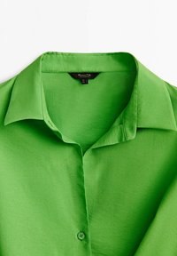 Chemise boutonnée vert vif à col, posée à plat, avec un col ouvert et une étiquette de marque visible à l'intérieur du col.
