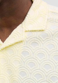 Camisa amarilla texturizada con un diseño de arcos superpuestos. Se pueden ver el cuello abotonado y el detalle de la tela suave.