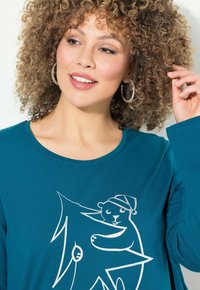 T-shirt mit langen Ärmeln in Teal, das ein weißes Grafikdesign eines Bären zeigt, der einen Weihnachtsmannhut trägt und einen sternförmigen Baum hält. Aus weichem Baumwollmaterial mit lockerer Passform.