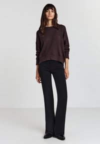 Maglione marrone a maniche lunghe con orlo a costine, abbinato a pantaloni neri a zampa d'elefante e scarpe nere basse, posizionato su uno sfondo neutro.