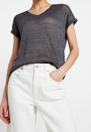 Personne portant un haut ample gris foncé à manches courtes et col en V, rentré dans un pantalon blanc taille haute, une main dans la poche.
