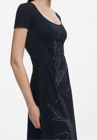 Robe noire avec des manches courtes, un décolleté en forme de cœur et un corsage ajusté. Motif graphique blanc remarquable avec des motifs botaniques sur le côté.