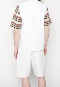 Weißes ärmelloses Oberteil, das über einem gestreiften Kurzarmhemd liegt. Die Shorts sind leicht und weiß, mit zwei Gesäßtaschen.