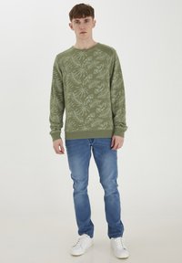 Sudadera verde oliva con un estampado de hojas de palma, cuello redondo y puños acanalados, combinada con jeans azules y zapatillas blancas.