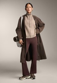 Długi brązowy płaszcz na beżowym swetrze na zamek, w połączeniu z bordowymi dopasowanymi spodniami i metalicznymi sneakersami. Pod jednym ramieniem trzyma zwiniętą matę do jogi.