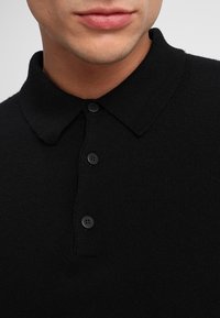 Svart stickad pikétröja med krage, som har tre svarta knappar och en texturerad tyg. Nära vy av kragen och knapparna.