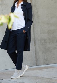 Marineblauer Trenchcoat über einem weißen Hemd, kombiniert mit marineblauen Hosen und hellgrauen Ankle Boots. Geschmeidiger Stoff und taillierte Passform sind bemerkenswerte Merkmale.