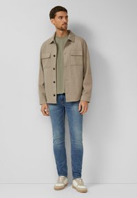 Beige overhemdjack met twee borstzakken, gedragen over een groen T-shirt, gecombineerd met een blauwe spijkerbroek en beige sneakers.