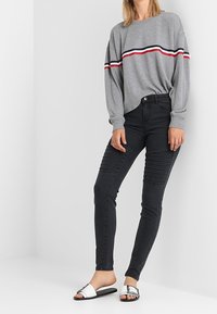 Šedý svetr s dlouhým rukávem, který má červené, bílé a námořnické pruhy, spárovaný s černými skinny džíny se strukturovanými akcenty a bílými skluznými sandály.