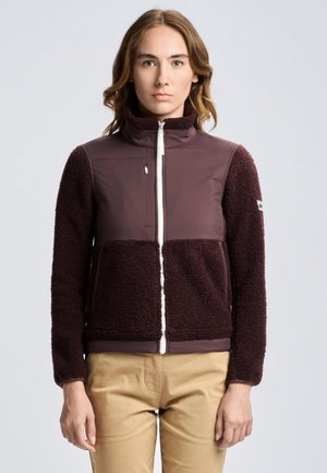 Una mujer luce una chaqueta de forro polar de color burdeos oscuro, con una cremallera delantera clara y pantalones de color tostado, sobre un fondo liso y claro.