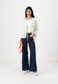 Grön- och vit-rutig väst med framfickor, tillsammans med mörka utsvängda jeans och vita sneakers. Håller en liten rosa handväska.