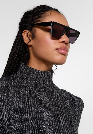 Donna con capelli intrecciati che indossa grandi occhiali da sole quadrati neri e un maglione dolcevita grigio scuro strutturato.